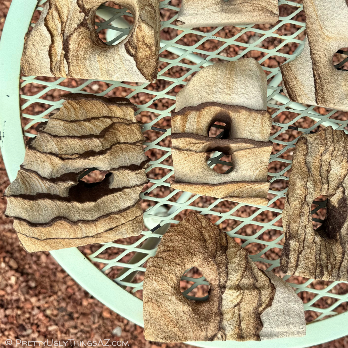 Light sandstone arches spread over a small mint-green table, with red gravel beneath them. They have pale tan color with faint to dark brown layering, and some have dark brown iron bands protruding.