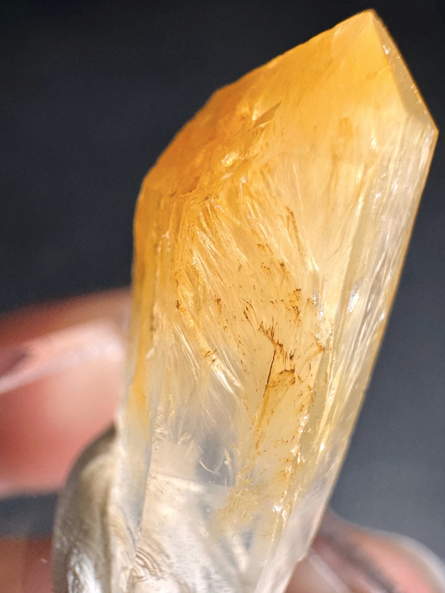 Close detail view of the halloysite fibers inside a quartz point, fine and hair-like parallel and curving fibers reflecting golden in the light, reaching from the surface of the tip into the quartz point in a gradient.