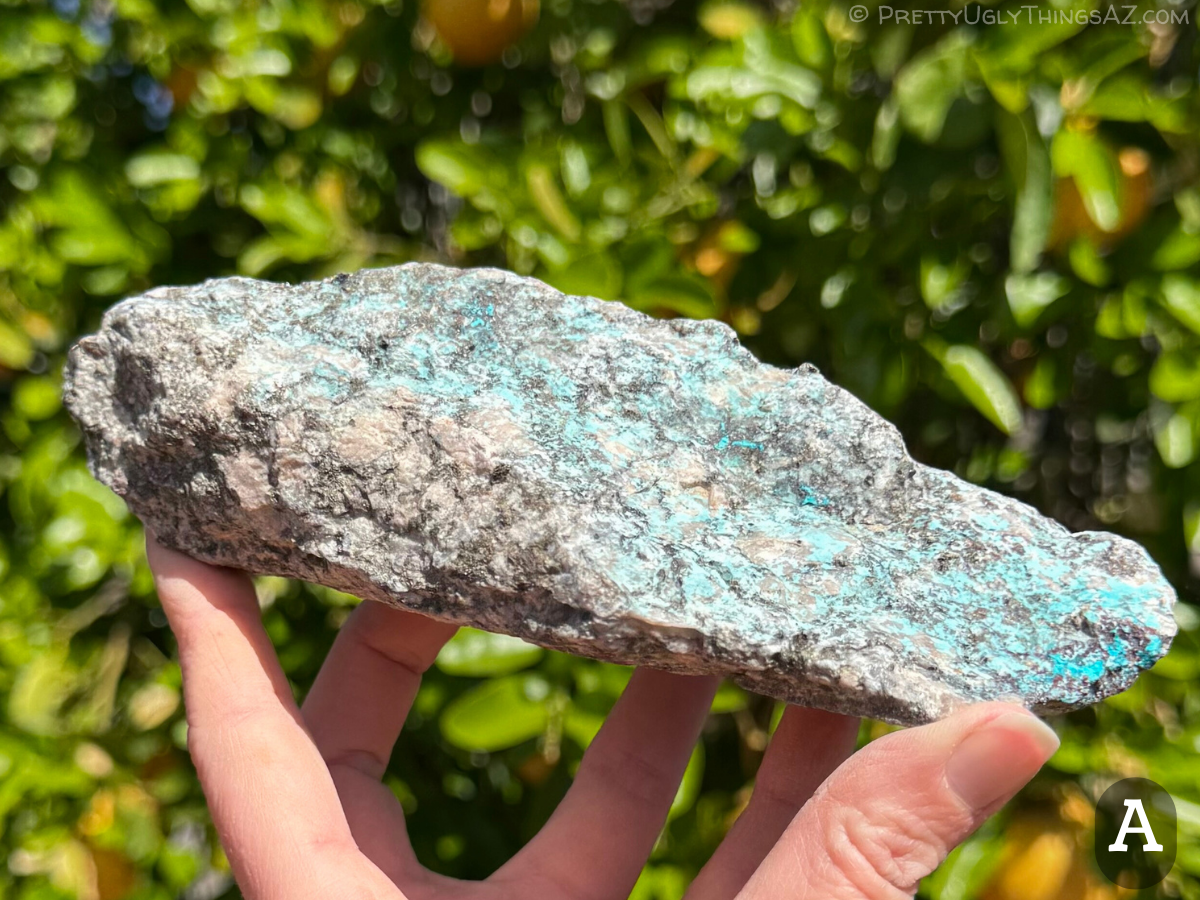 Option A, a long narrow rough piece of light-colored granite with black speckles and some chunky feldspar crystals visible. The top has a light blue mineral coating, with vivid blue chrysocolla on the near end.