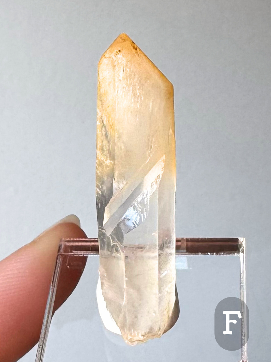 Option F, a long narrow point with pale orange-yellow coloration in the tip, which is milky and semi-transluscent. The inclusions catch light well, making the point bright. The point has a diagonal impression left from a small point that once grew alongside it.