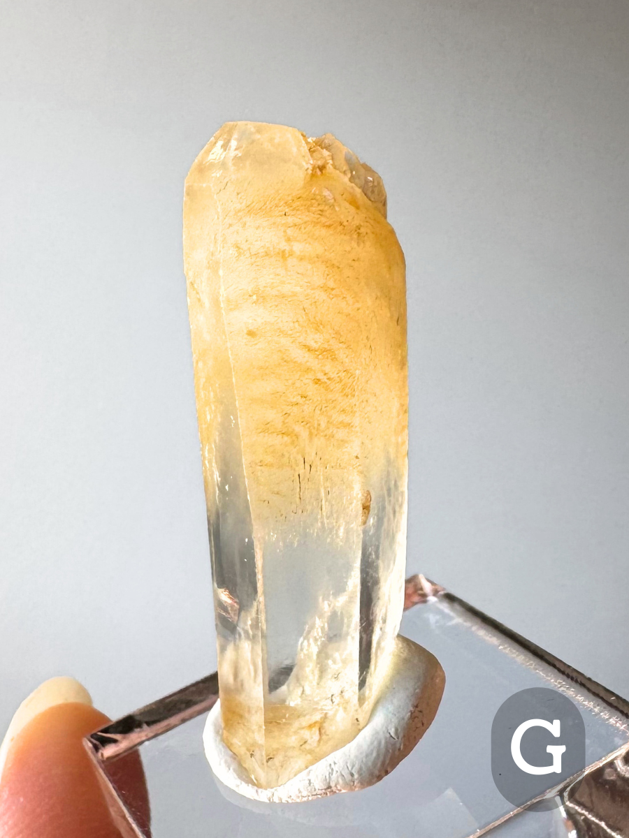 Option G, a long thick quartz point with an odd drill-bit style termination. Orange-yellow halloysite inclusions fill the top half, with a slightly wavy appearance with chatoyancy like hair. The base is clear.