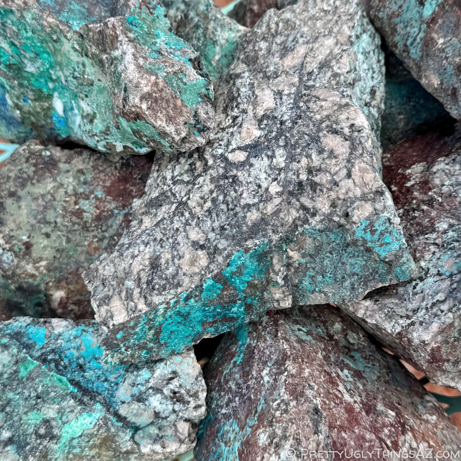 A pile of rough rocks with turquoise-blue color running through them. The rocks are a mix of colors, some dark brown and some near-white and speckled with black spots and large light-colored feldspar crystals.