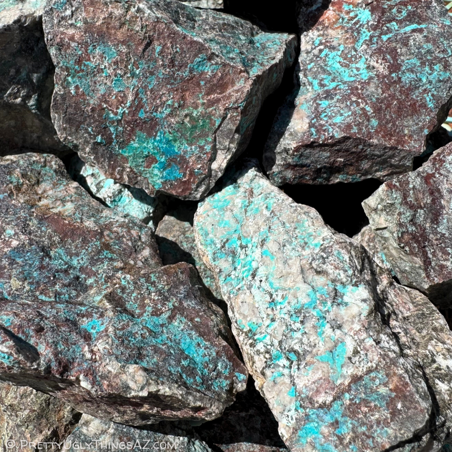 A pile of rough rocks with turquoise-blue color running through them. The rocks are a mix of colors, some dark brown and some near-white and speckled with black spots and large light-colored feldspar crystals.