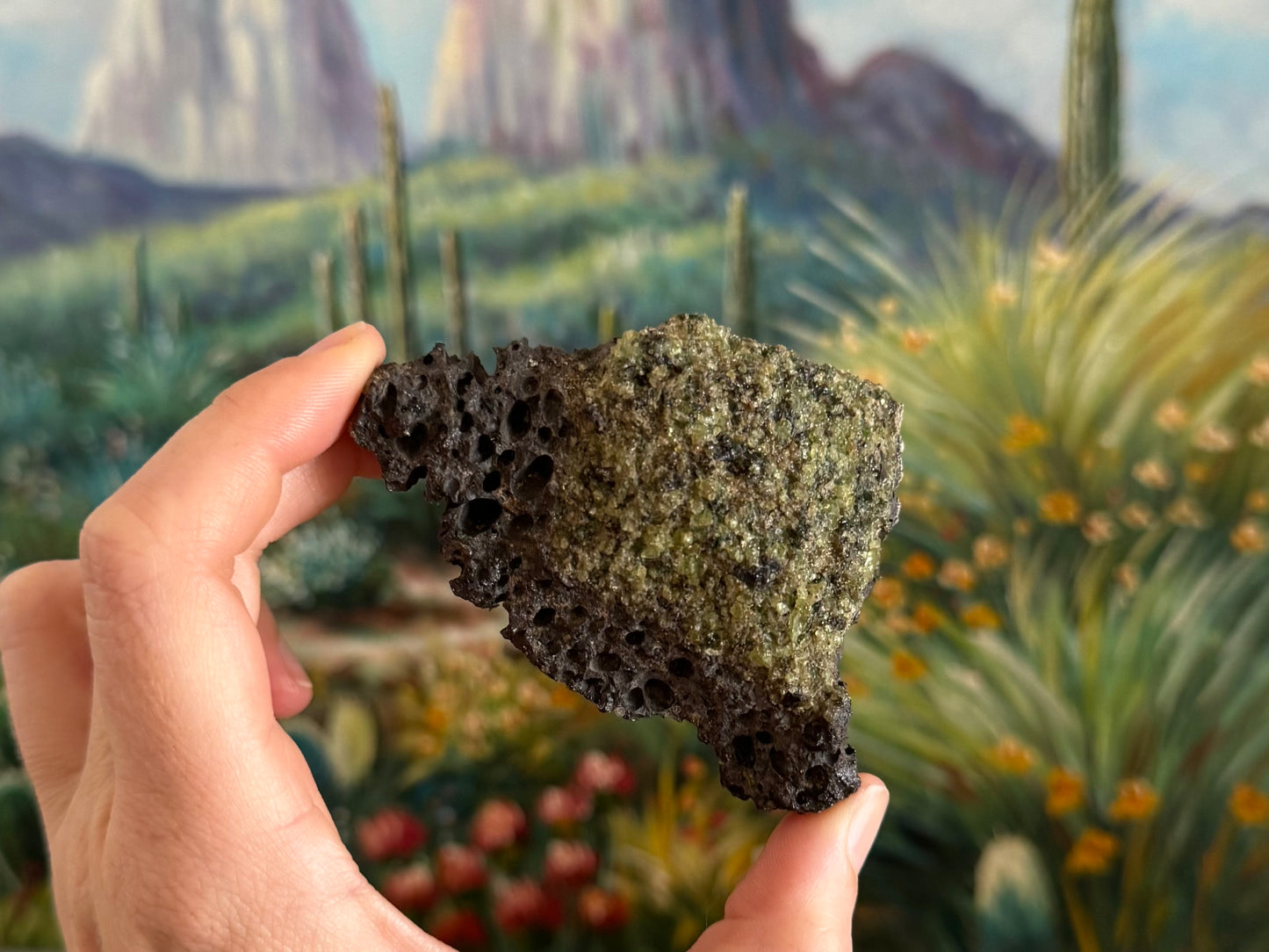 A rough triangular chunk of dark gray porous volcanic rock with large bubble holes. There is a patch of low quality peridotite covering most of the front, with desaturated green color and poor luster and clarity.