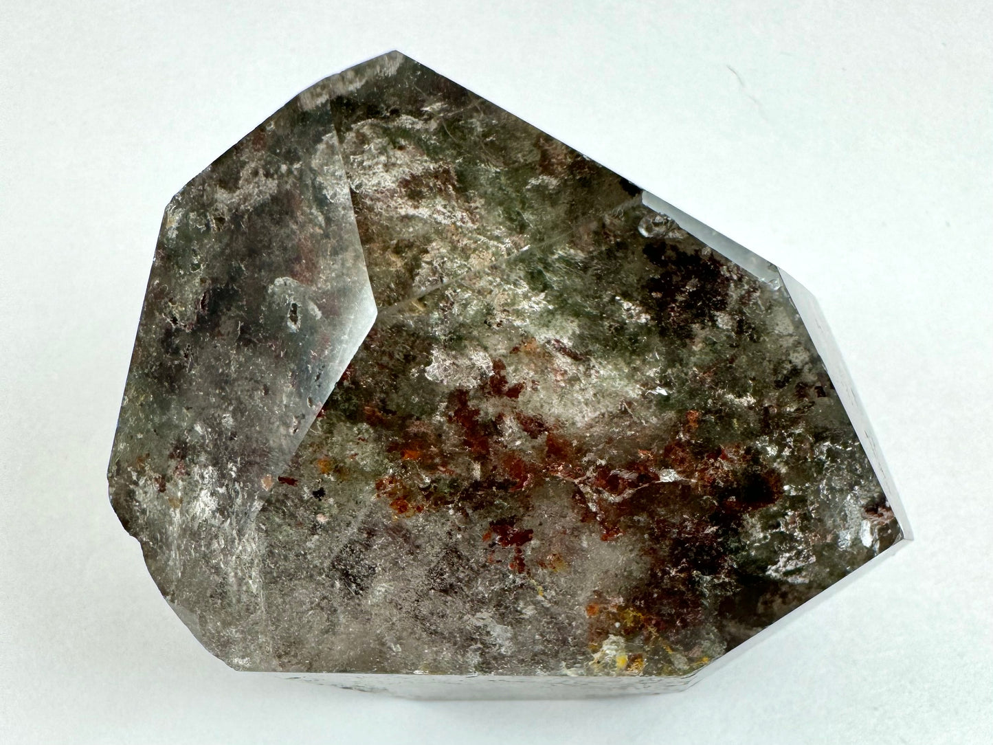 Close view of a piece of garden quartz shaped like an angular shield and perfectly colorless to show green, white, and red-brown chlorite inclusions inside. The surface has high luster and where it reflects small indentations are visible on the surface.
