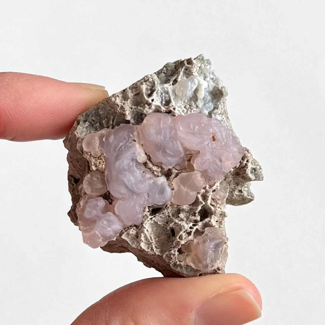An off-square piece of light gray rock matrix with a triangular cluster of light pink small knobby botryoidal forms covering the middle third of the piece. There is more chalcedony with light yellow-pink tint in the bottom corner, and some white low-quality chalcedony in the upper right.