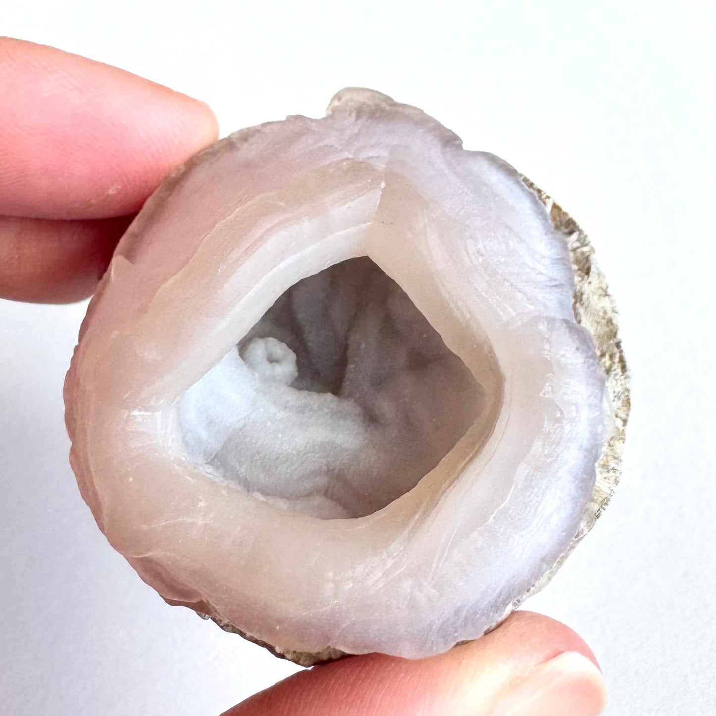 A pink chalcedony nodule with light pink color, near-round with ropy layers making a cup-nest shape. The concentric banding is smaller around a wonky opening in the top with a hollow interior. Inside the center, there is a small white raised ring with more subtle undulating banding radiating out around it, all with very fine druzy texture.