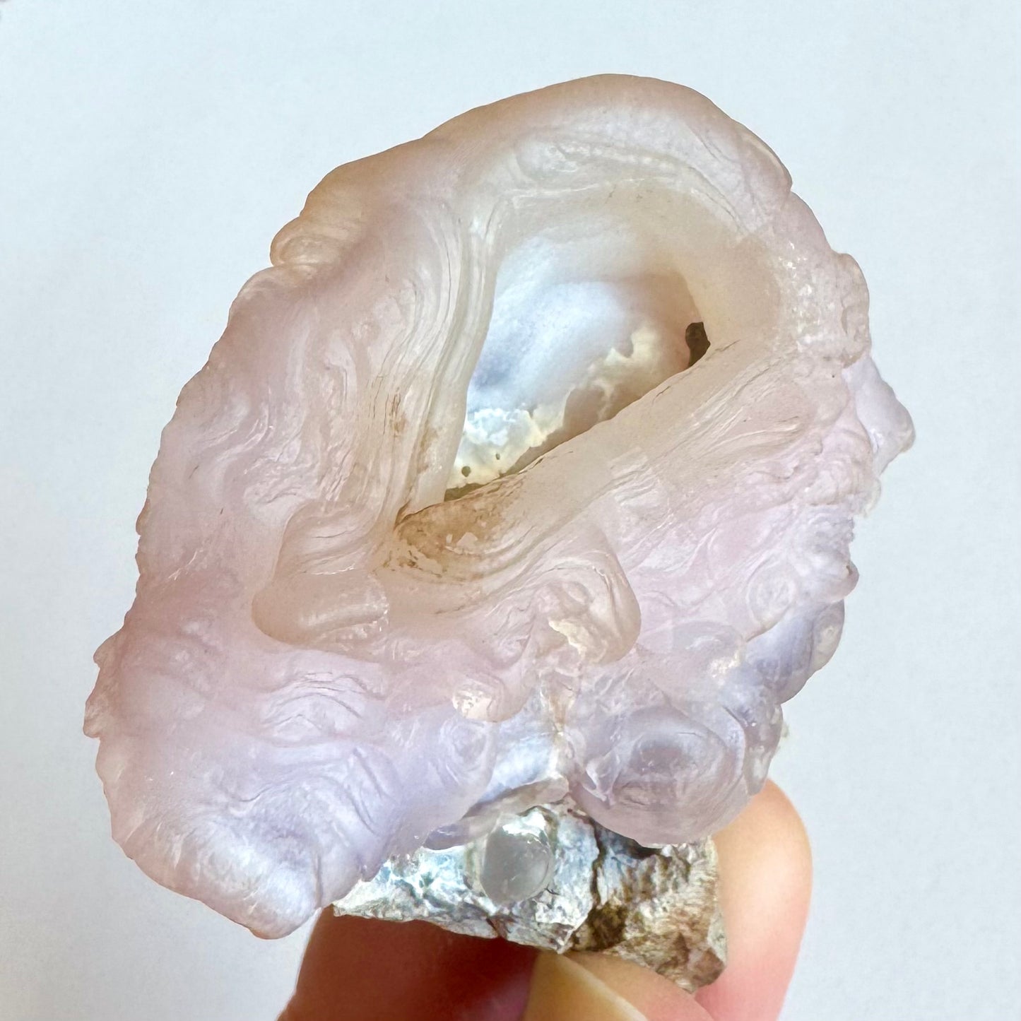 An elongated pink chalcedony nodule with a small angular opening around a hollow interior, fluid ropy banding in concentric rings down the sides. The swirling layers quickly become blobby botryoidal forms around the sides. There is a dark piece of rock lodged deep inside the interior.