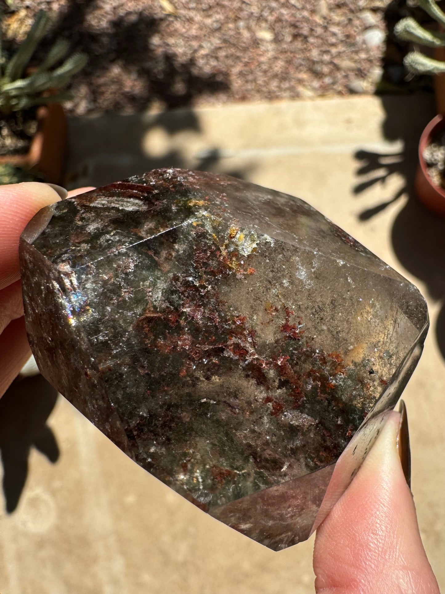 The piece held down with light shining on it, with brick-red and green inclusions visible inside and some clarity near the top right. There is a rainbow in a small fracture on the left side.