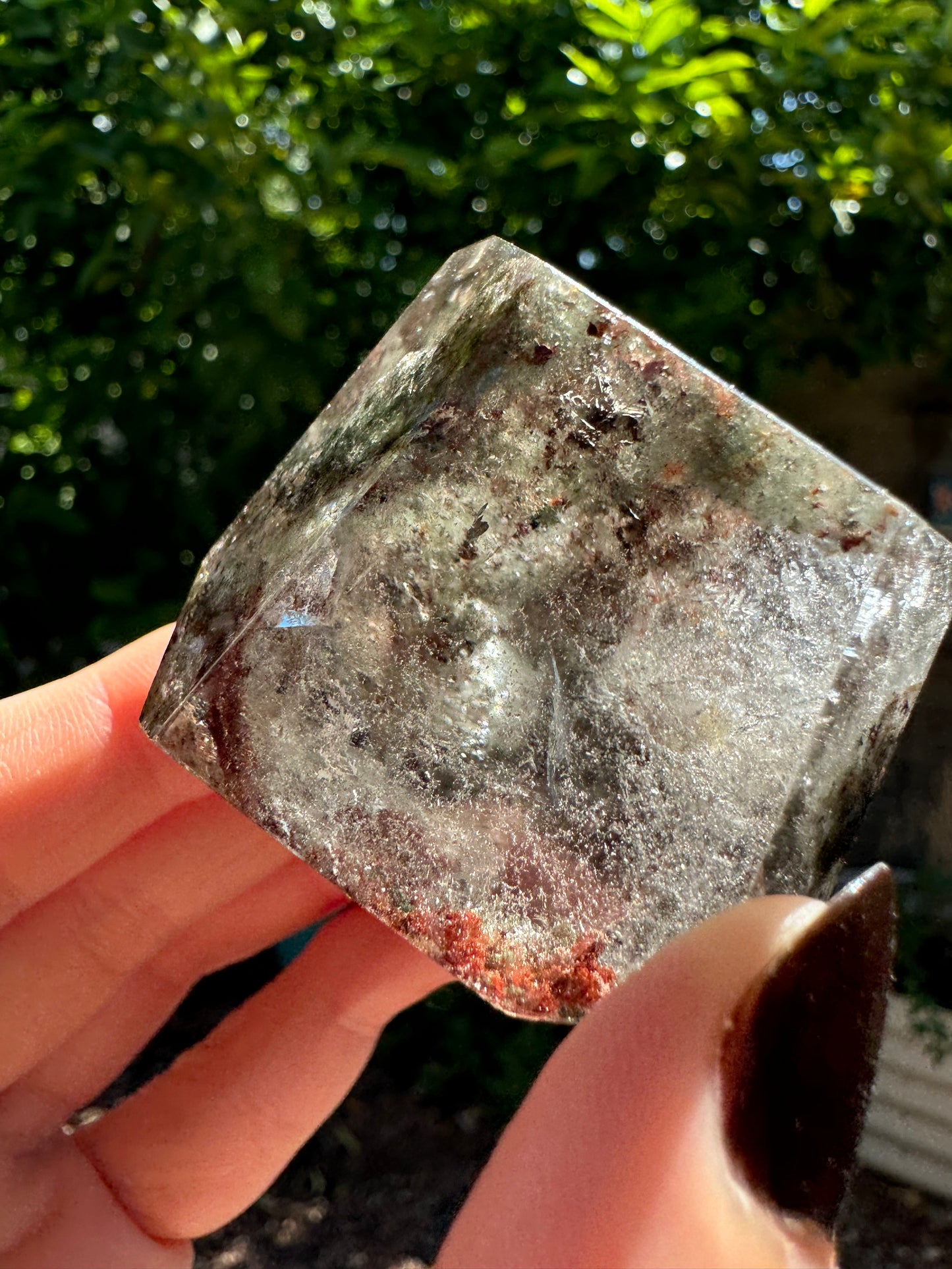 Another end of the quartz piece lit by sun from above, with green inclusions along the top left, and bright red ones at the bottom. Wispy inclusions or illuminated throughout the piece.