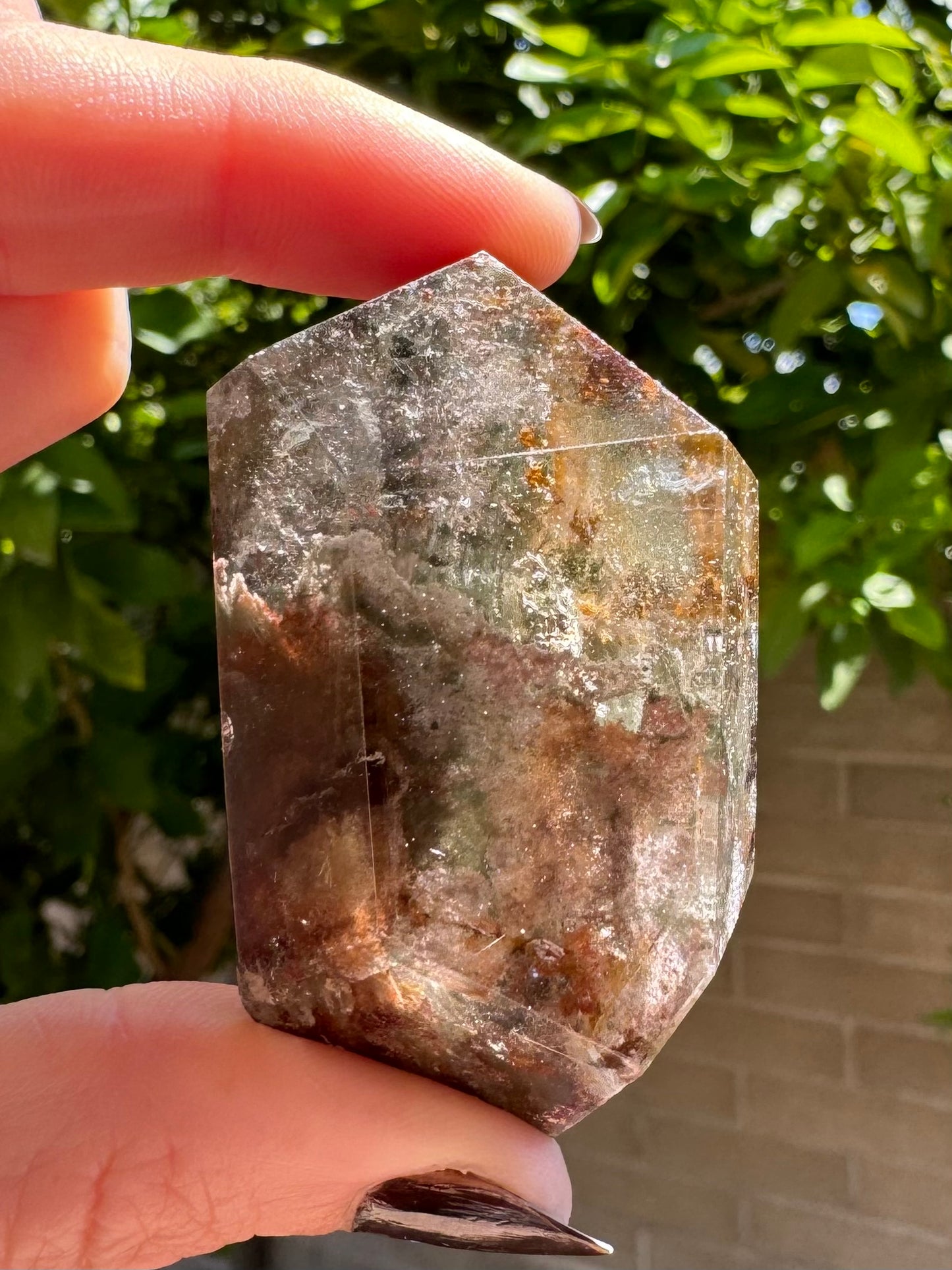 The lodolite specimen held between thumb and finger for scale, in sunlight to highlight internal features. The freeform cut is barrel-shaped, pointed on each end.