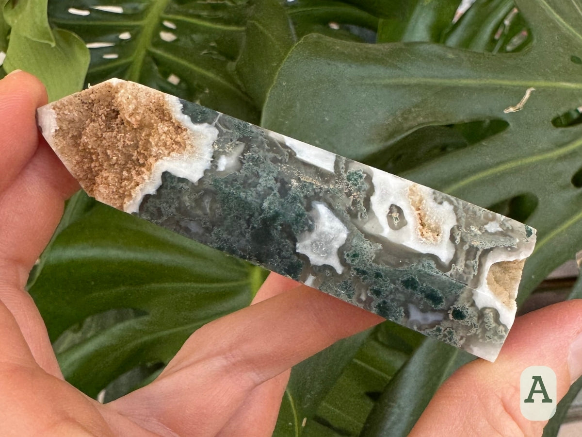 Option A, a six-sided moss agate tower with mossy blue-green inclusions swirling through clear and white chalcedony. The tip is missing a side, which is a rough cavity