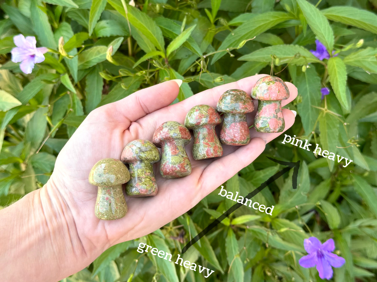 Hand with six unakite mushroom lined up across it. It is labeled "green heavy" at the left end, "balanced" in the middle, and "pink heavy" at the right end. The mushrooms are colored accordingly: on the left they are mostly green with small pink spots; in the center green with larger pink spots; on the right green covered in large pink spots