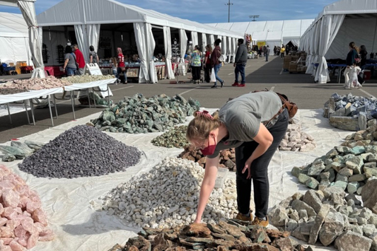 Mineral show with person bending over to look at a pile of rocks among other piles of rocks, and booths in the background