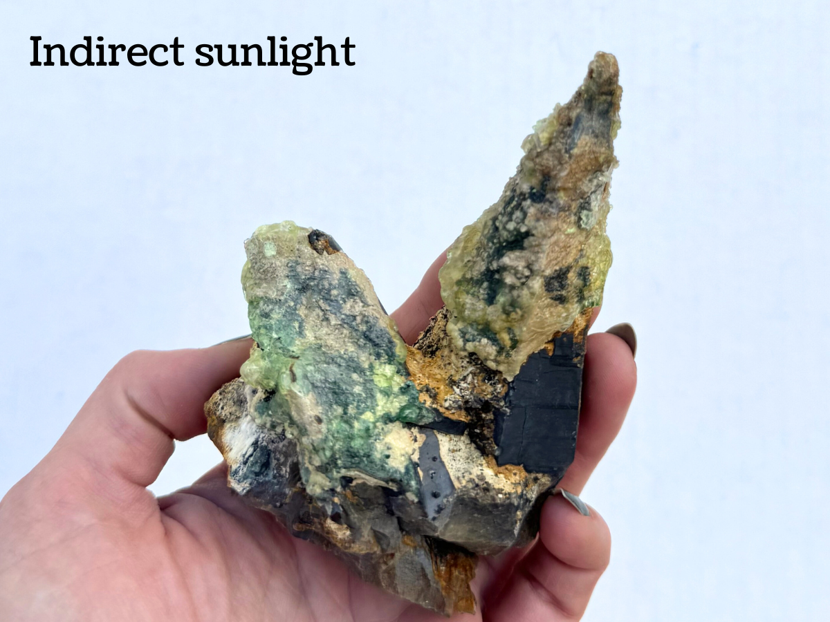 The piece held in a hand for scale, partially filling the hand. It is captioned "indirect sunlight" and the hyalite is glowing a light green color.