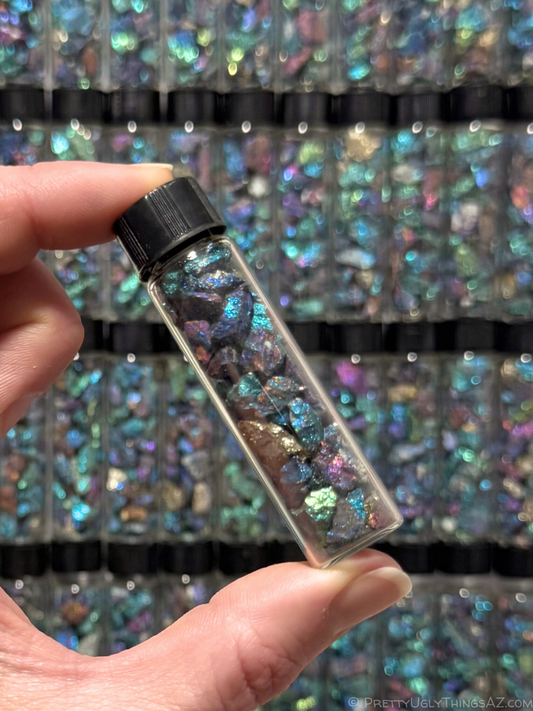 A vial of small irregular chips of iridescent chalcopyrite, held between thumb and forefinger. In the background there are stacked rows of more of the vials. The vial is long and slender with a black plastic lid. The chalcopyrite is a mix of blue, purple, green, and a cool gold, with some flashes of orange.