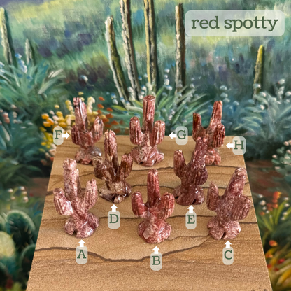 Captioned "red spotty," eight tiny identically carved saguaros on a piece of sandstone labeled A through H. A is a red-white with a dark red vein and spotting. B is bright red with light patches. C is a grainy desaturated dark red and white. D has bold dark brown-red and off-white pattern. E looks like salami, dark red with light red speckles. F is brick red with light splotches. G is dark red with streaky lighter color. H is a deep red with light spots.