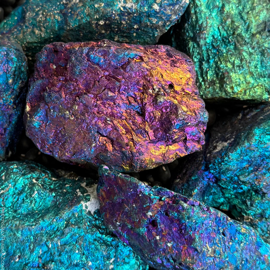 Close view of a pile of rough chunks of chalcopyrite with different iridescent colors. The one in the focus at center is pink-purple with orange and blue flashes. The pieces around it have a mix of yellow-green, blue-green, blue, violet, and purple iridescence.