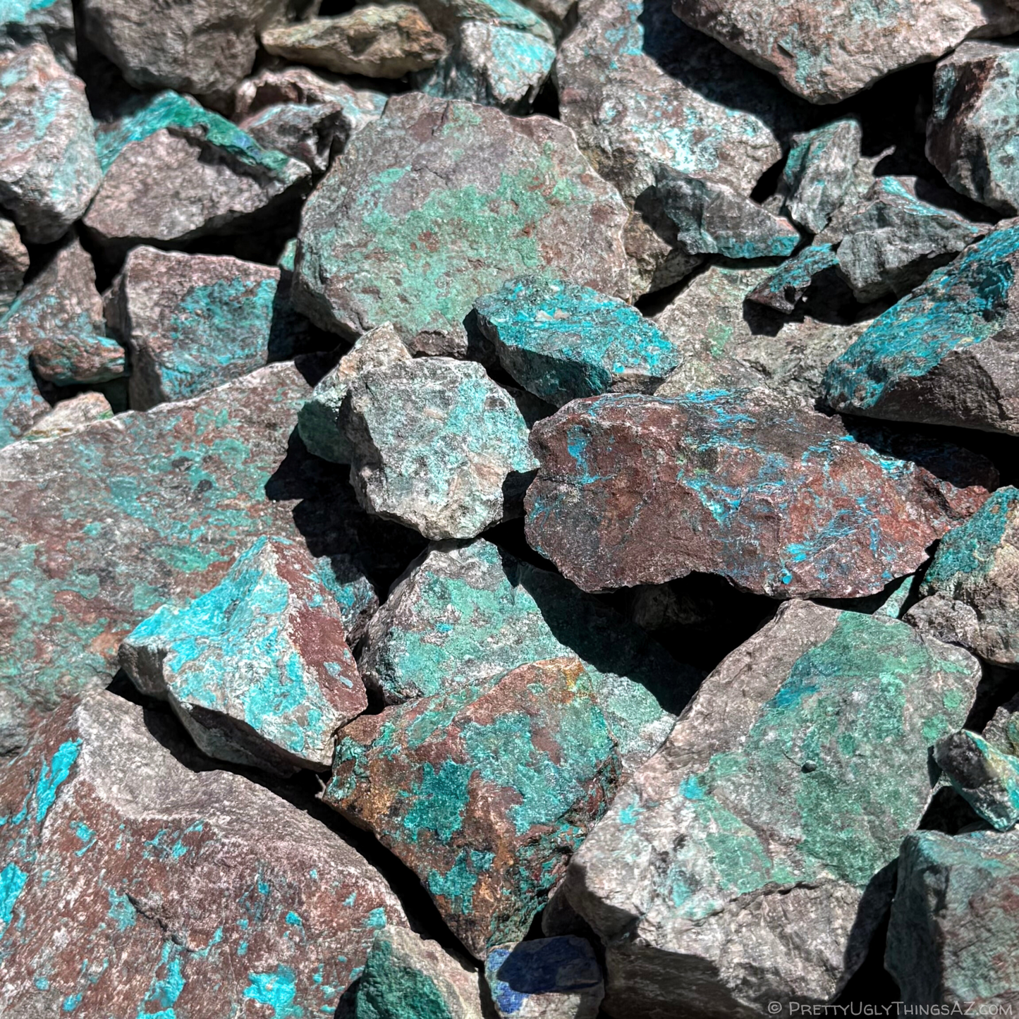 A pile of rocks with patches of varying shades of aqua blue and green copper minerals, the bright color contrasting with dark red-brown and patchy salt-and-pepper host rocks.