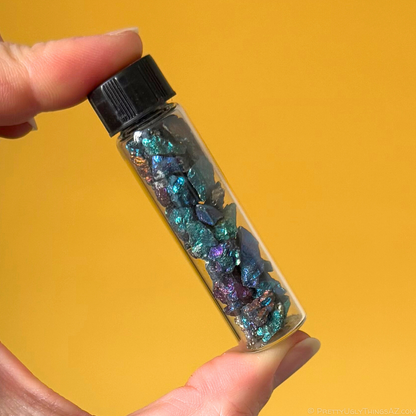 One of the chalcopyrite vials held in front of a contrasting bright yellow background. The material is fairly dark but with strong peacock tones of green, blue, purple, and orange.