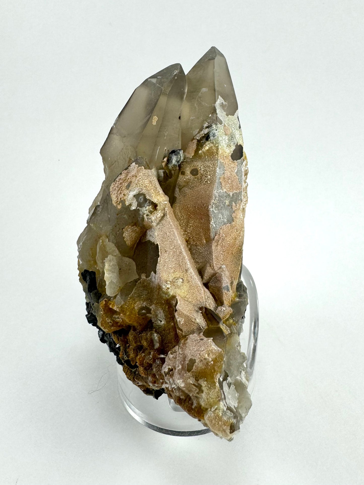 The first of three lighting images: two large smoky quartz crystals, appearing intergrown as possible twins, with parts of several partial quartz crystals below at a slant. The bottom and right side of the specimen is covered in a thin hyalite crust with opaque light brown coloration under it. The hyalite has cheese holes in it.