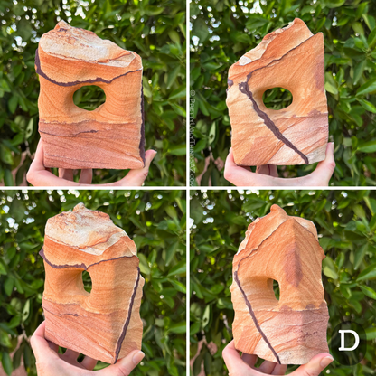 Option D, a blocky squat rectangular arch with a rough slanted top. The piece has color divided in three sections like candy corn, the bottom third a purple-tinted orange, the middle orange, and the top near-white. A couple angular bold dark iron bands cut across the faces. The sandstone has subtle layering.