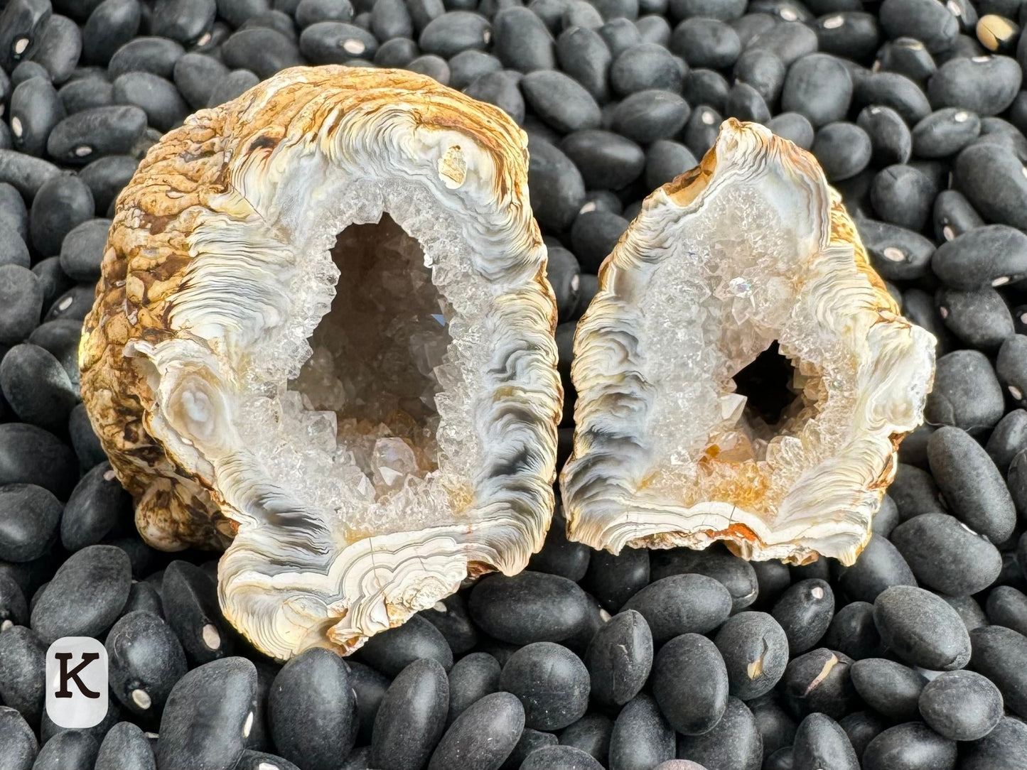 Option K: an off-round geode pair with one half larger than the other. The interiors are lined with colorless quartz which turns into white chalcedony streaked with swirls of darker brown and yellow-tan.