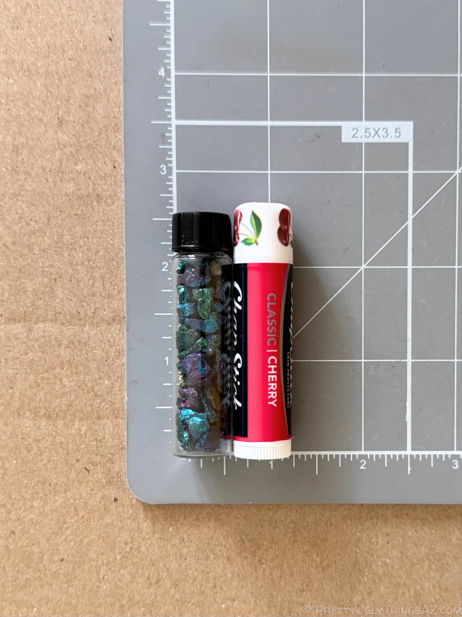 A vial on a measuring mat for scale, measuring barely under two and a half inches long and about half an inch wide. There is a chapstick placed next to it to show that it is almost the same size, but slightly larger than the vial.