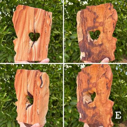 Option E, a broad flat arch shaped like a tablet with an angled top. A thin dark iron band runs down the middle of the face, making the hole in the middle heart-shaped. One face of the arch has excellent high-contrast layering, with deep red-orange to light cream layers. The other face is nearly totally covered with dark iron banding in a flat layer on the surface,  blotchy dark brown to orange-red color.