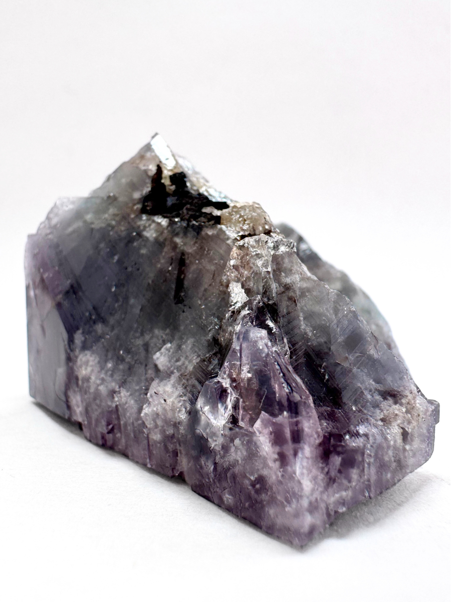 The specimen face-down, the pointed broken backside sticking up like a mountain range with black iron oxides attached along the ridge. The surface is purple, while the interior is faintly green tinted.