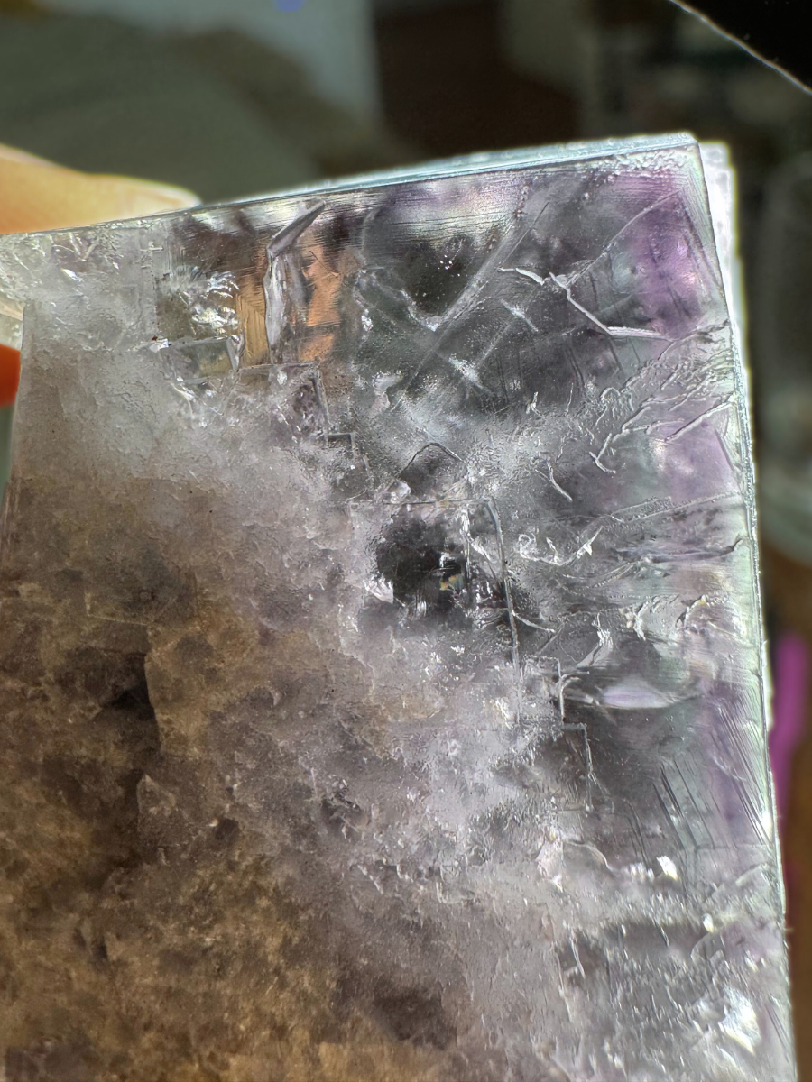 A close detail view of a corner of the piece brightly side-lit to emphasize the color zoning. There are thin concentric square bands light green on the surface, purple, then a fainter alternating layers inside. The white inclusions inside look fuzzy and near-dendritic.