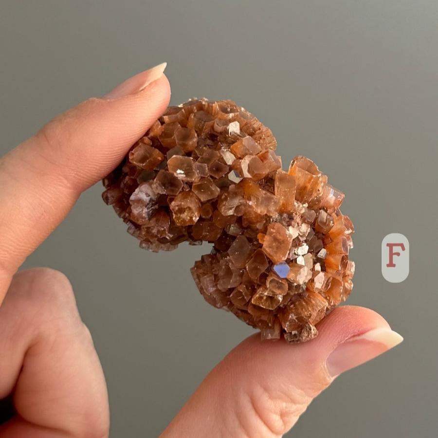 A sputnik aragonite cluster shaped like two spheres, labeled 'F,' held in the fingers to show small scale.