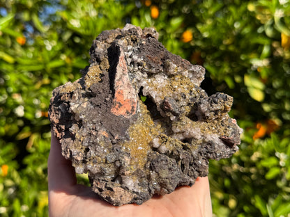 A large specimen held in a hand, approximately the size of two fists. The piece is chaotic, dark iron oxides form a craggy surface with many pockets and folds, and a wedge of orange-tinted rock matrix is at the center, next to a small hole where you can see through the center of the specimen. There are patches of a golden yellow and a colorless mineral on the piece, interspersed with the red rock and near-black matrix.
