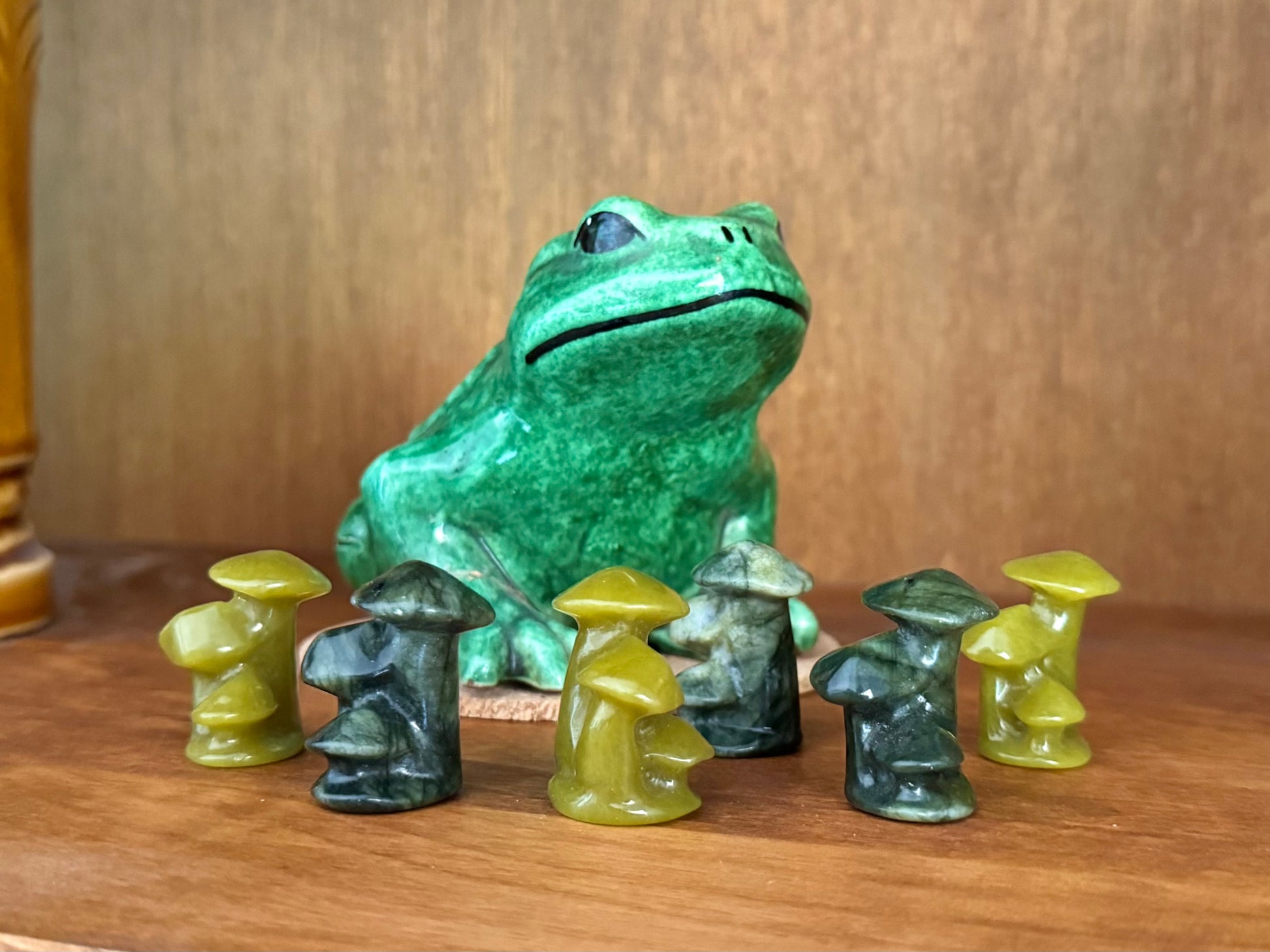 Several carved mushrooms in yellow green and forest green minerals, trio clusters of three mushrooms of different heights. They are arranged on a wood shelf in front of a ceramic frog.