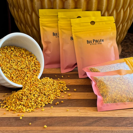 A pile of sealed pouches labeled "Bee Pollen 0.75 oz / 22 g" with granular yellow pieces visible through the window. To the left a small white bowl is spilling bee pollen granules.