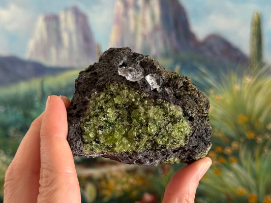 A triangular rough chunk of dark gray porous volcanic basalt with a large patch of peridotite on the front face and a patch of darker crystals on the upper right.