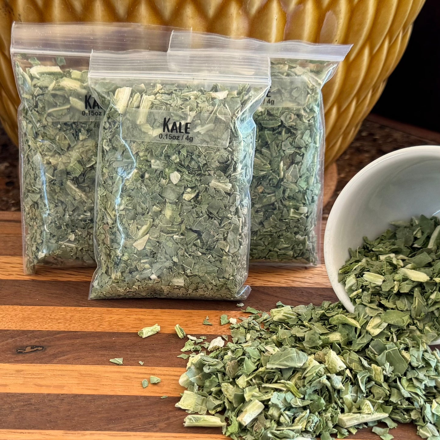 Several bags filled with kale flakes, labeled "Kale 0.15oz / 4g," with a small bowl to the right spilling dried kale flakes