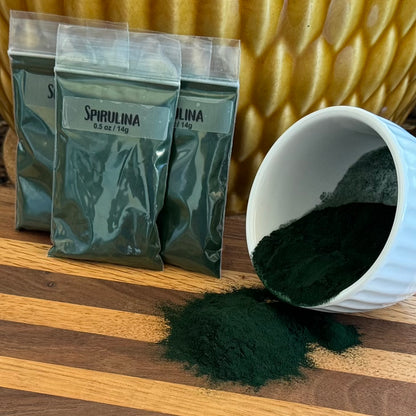 A pile of ziploc bags labeled "Spirulina 0.5 oz / 14g" filled with dark blue-green powder. To the right a small white bowl is spilling spirulina powder.