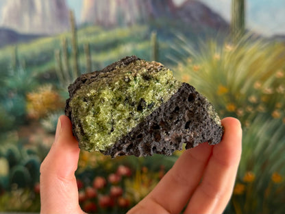 A rough irregular chunk of dark gray porous volcanic basalt with bubble holes. There is a patch of gemmy peridotite across the left side of the face with dark crystals in the mix.