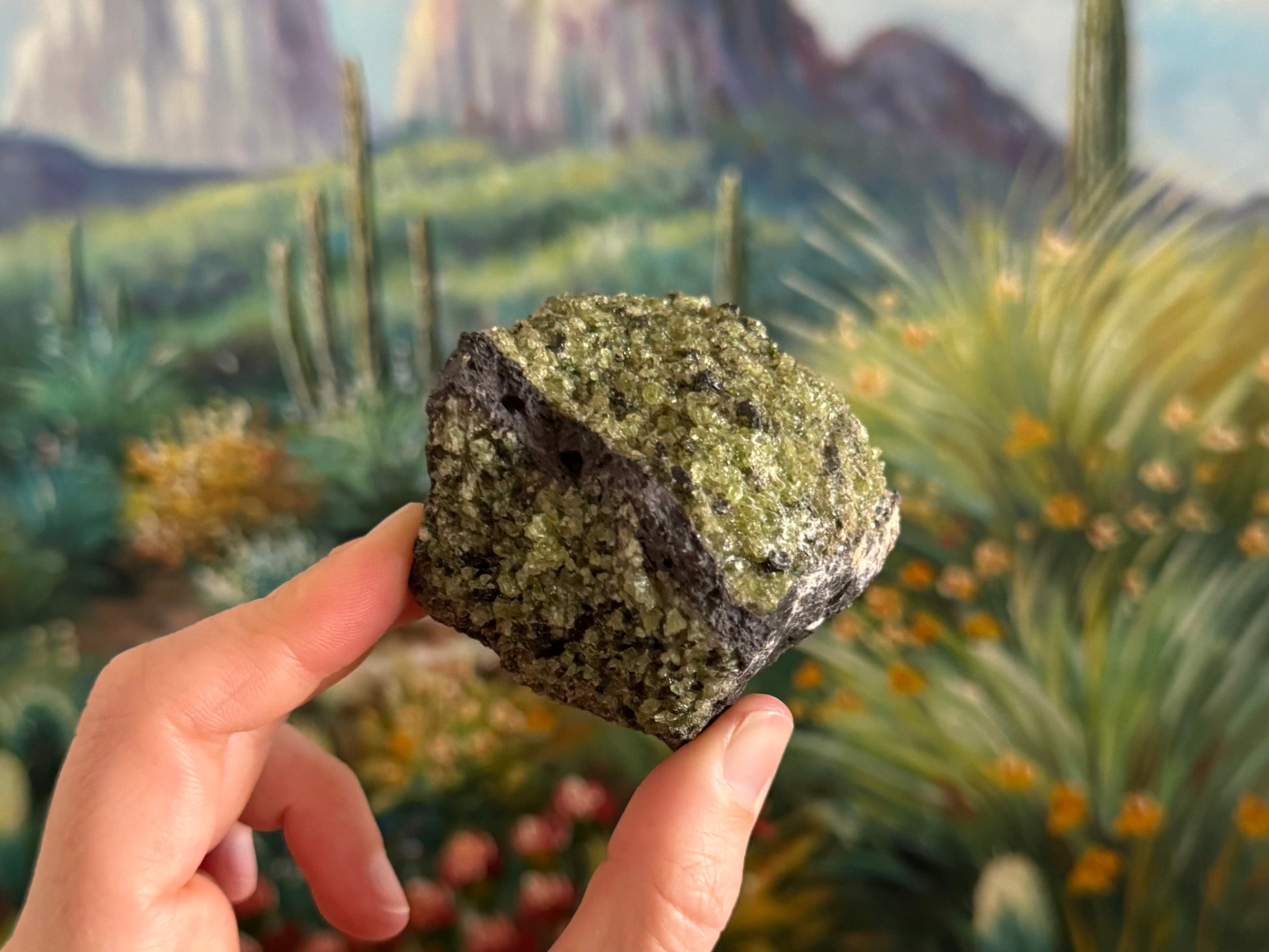 A rough compact chunk of dark gray porous volcanic basalt with bubble holes and white overgrowth in areas. There is a patch of gemmy peridotite across the top and front faces with dark crystals in the mix.