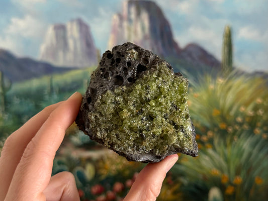 A rough irregular chunk of dark gray porous volcanic basalt with bubble holes. There is a patch of gemmy olivine-heavy peridotite on the front face with some dark crystals in the mix.