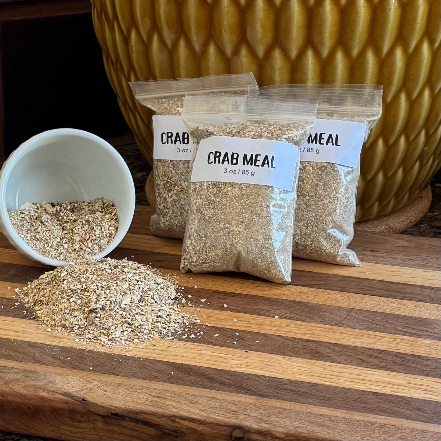 A pile of ziploc bags labeled "Crab Meal 3 oz / 85g" filled with fine grain white and tan pieces. To the left a small white bowl is spilling crab meal