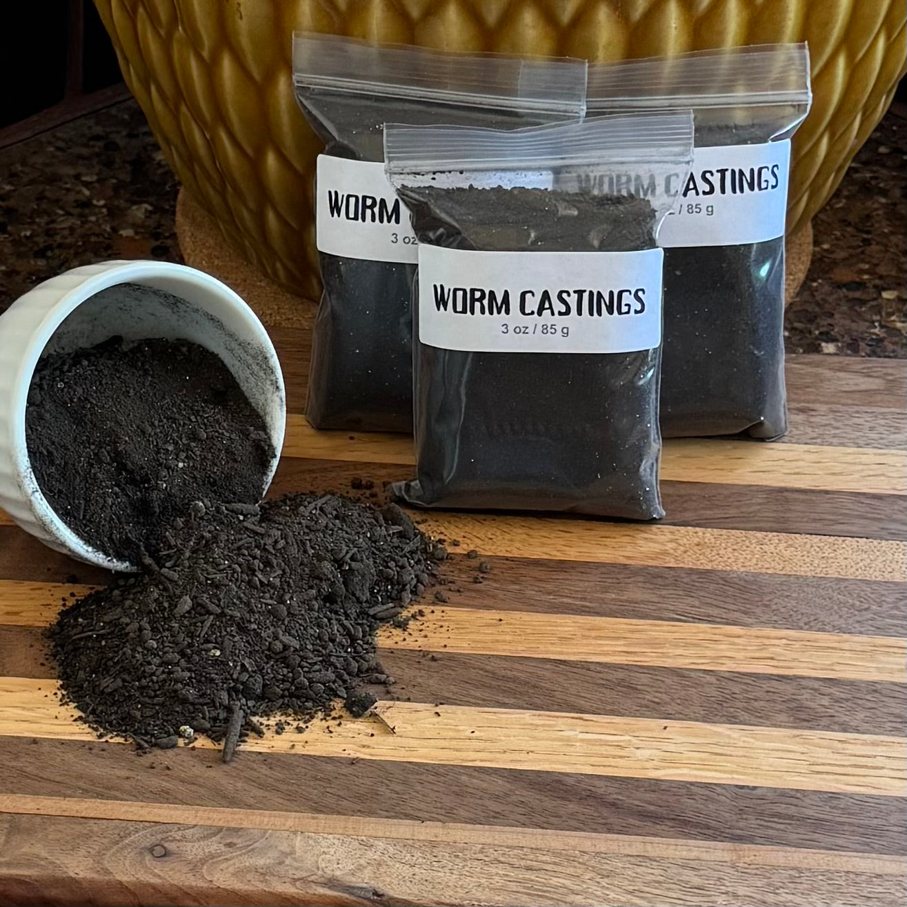 A pile of ziploc bags labeled "Worm Castings 3 oz / 85g" filled with dark soil. To the left a small white bowl is spilling worm casting that look like soil.