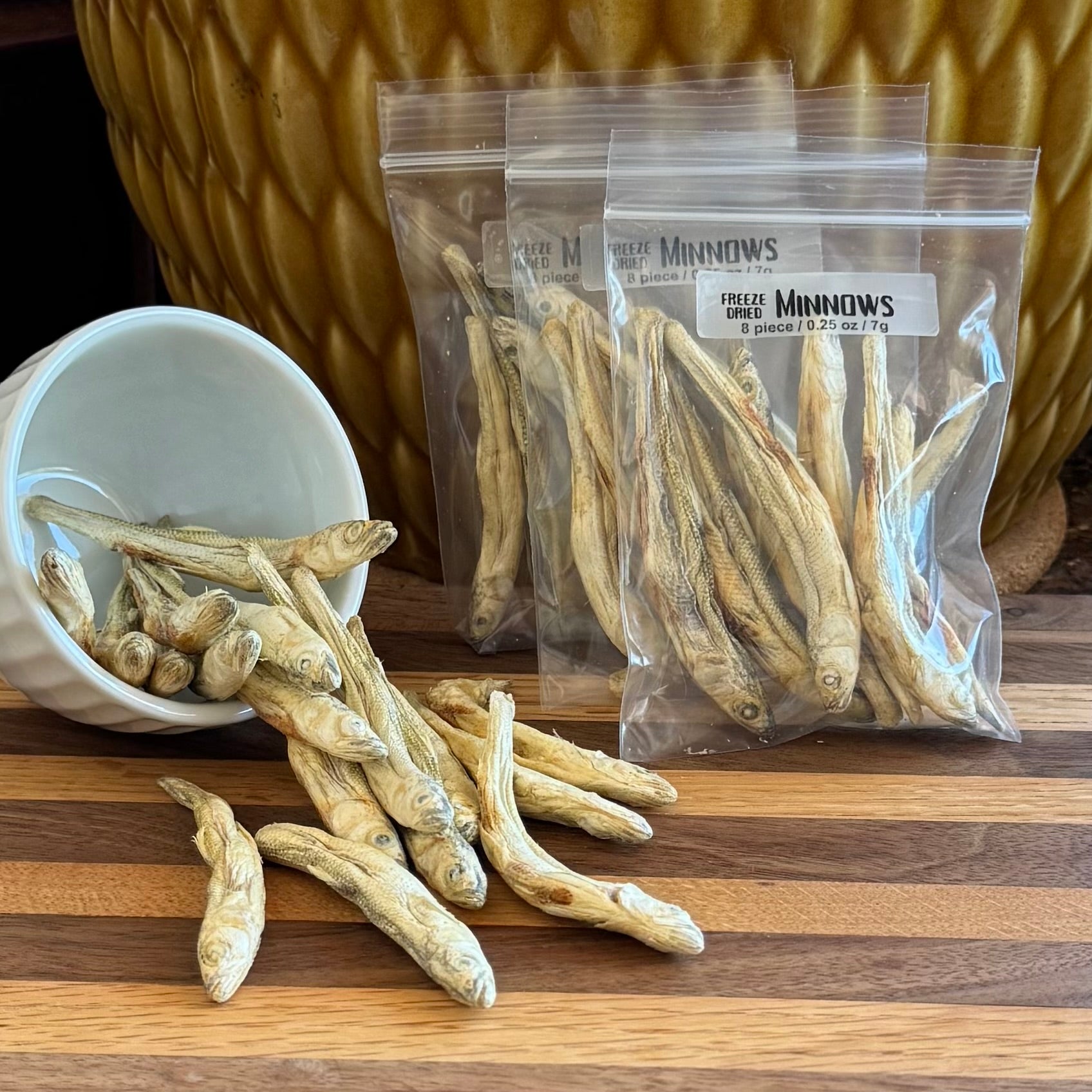 Several bags filled with dried minnows, labeled "Freeze-dried Minnows 8 piece / 0.25oz / 7g," with a small bowl to the left spilling dried minnows