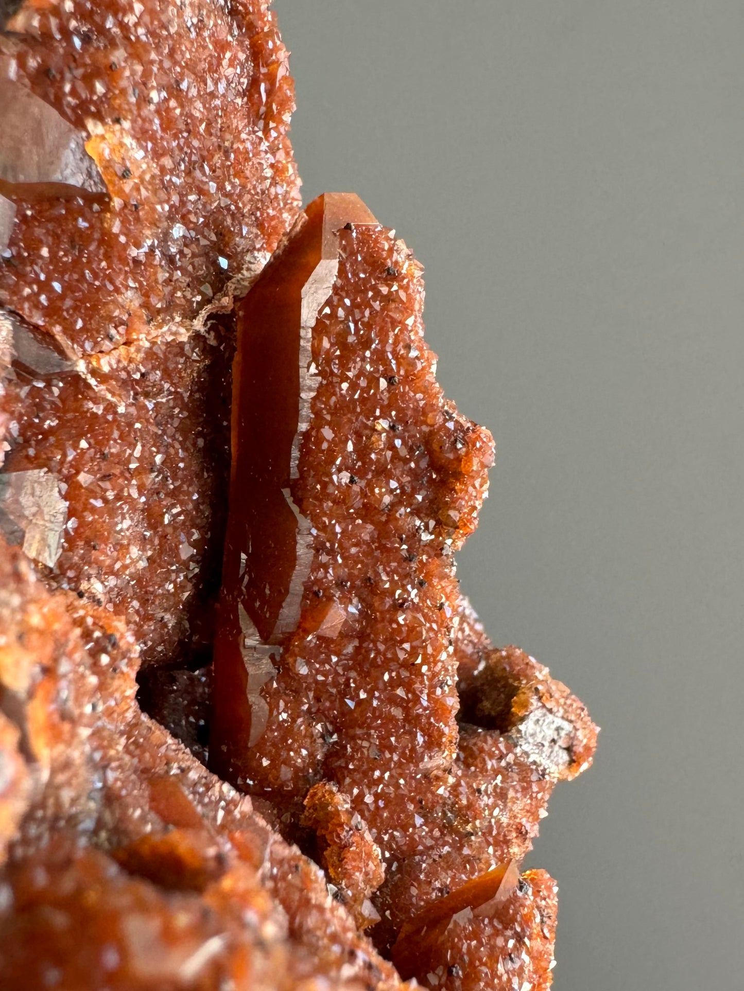 Detail of a quartz point on the specimen in indirect light, with excellent form visible on one side and the other side covered with thick glittery red druzy with black dots.