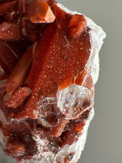 Detail of several hexagonal broken crystals with colorless interiors. The side of a wide red quartz crystal above is covered with red druzy with black spots.