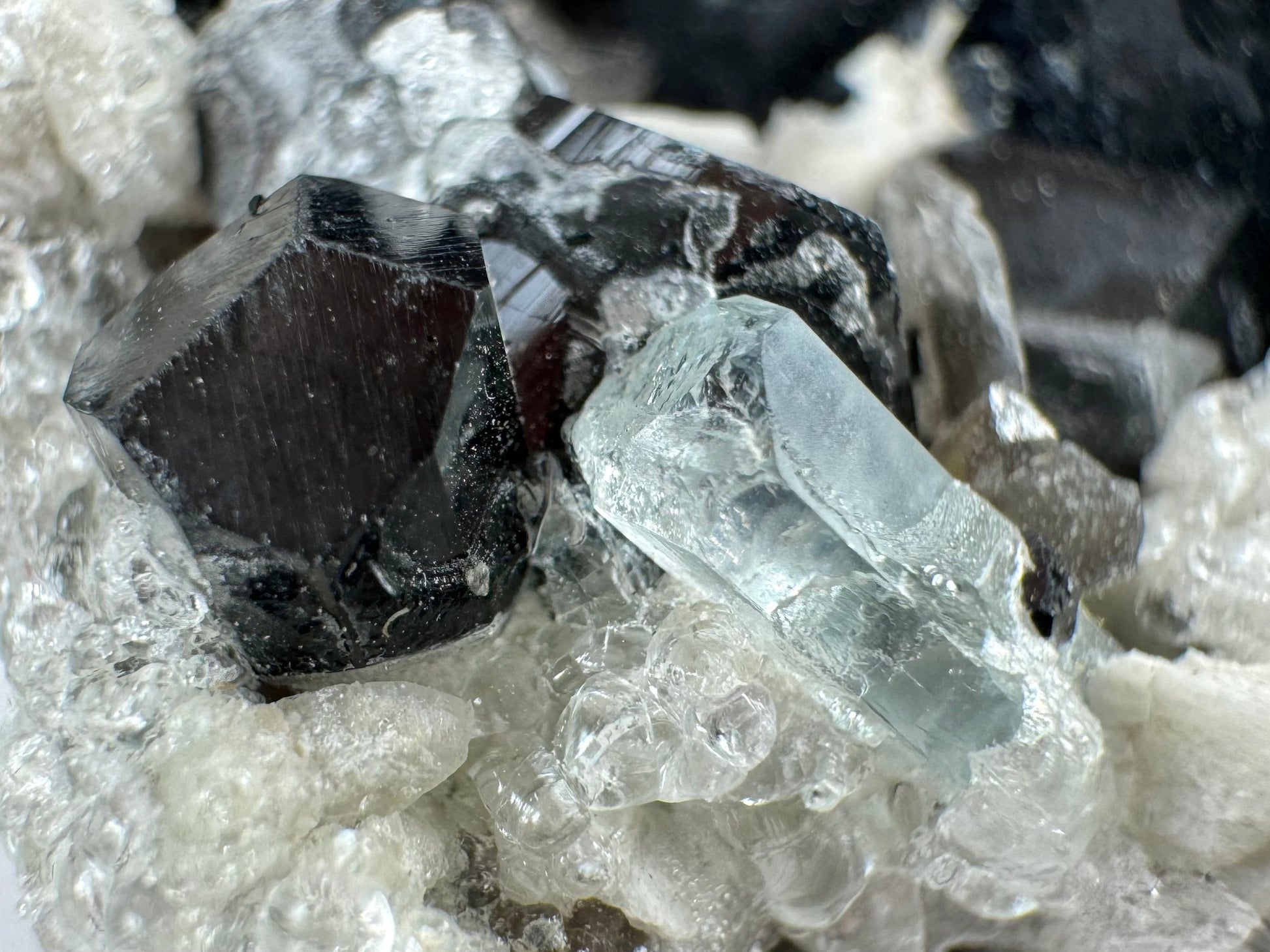 Detail of the small aquamarine crystal, a six-sided prism with light blue coloration. It is partially embedded in hyalite, with large spheres of hyalite forming nearby. A well-formed small schorl crystal is to its left.