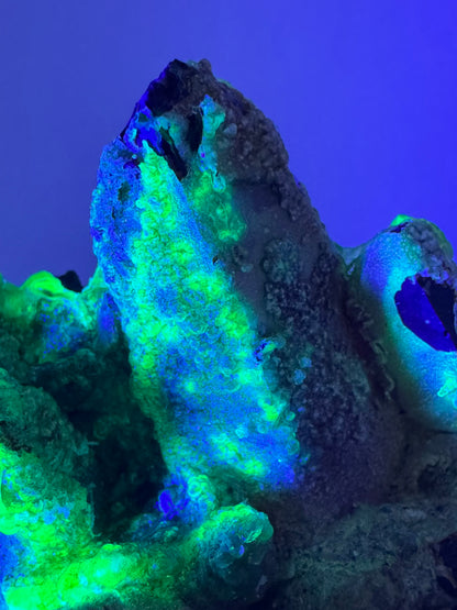 Detail of a smoky quartz crystal coated in hyalite, in the dark illuminated with UV light. It has patchy bright yellow-green and icy blue coloration, with spherical growth visible.