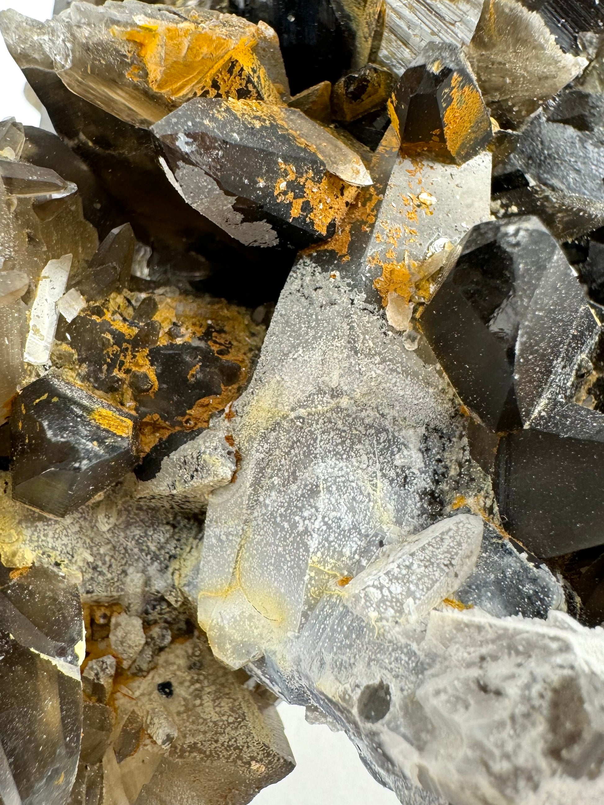 Detail of the overgrowth on the surface of several smoky quartz prisms in the cluster. The rust-orange color is opaque and rough. The white appears like a milky translucent film with round white spots throughout