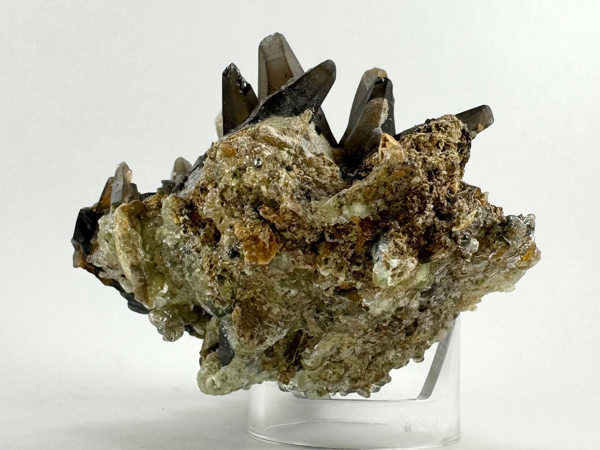 The second of two lighting images: the specimen in the same position as the previous image, in white light. The points are dark-colored, and the bottom is coated in minerals. There is a light brown aggregate of irregular growth, with shard-like pieces of smoky quartz and iron staining. It is covered in a slightly green-yellow layer of hyalite.